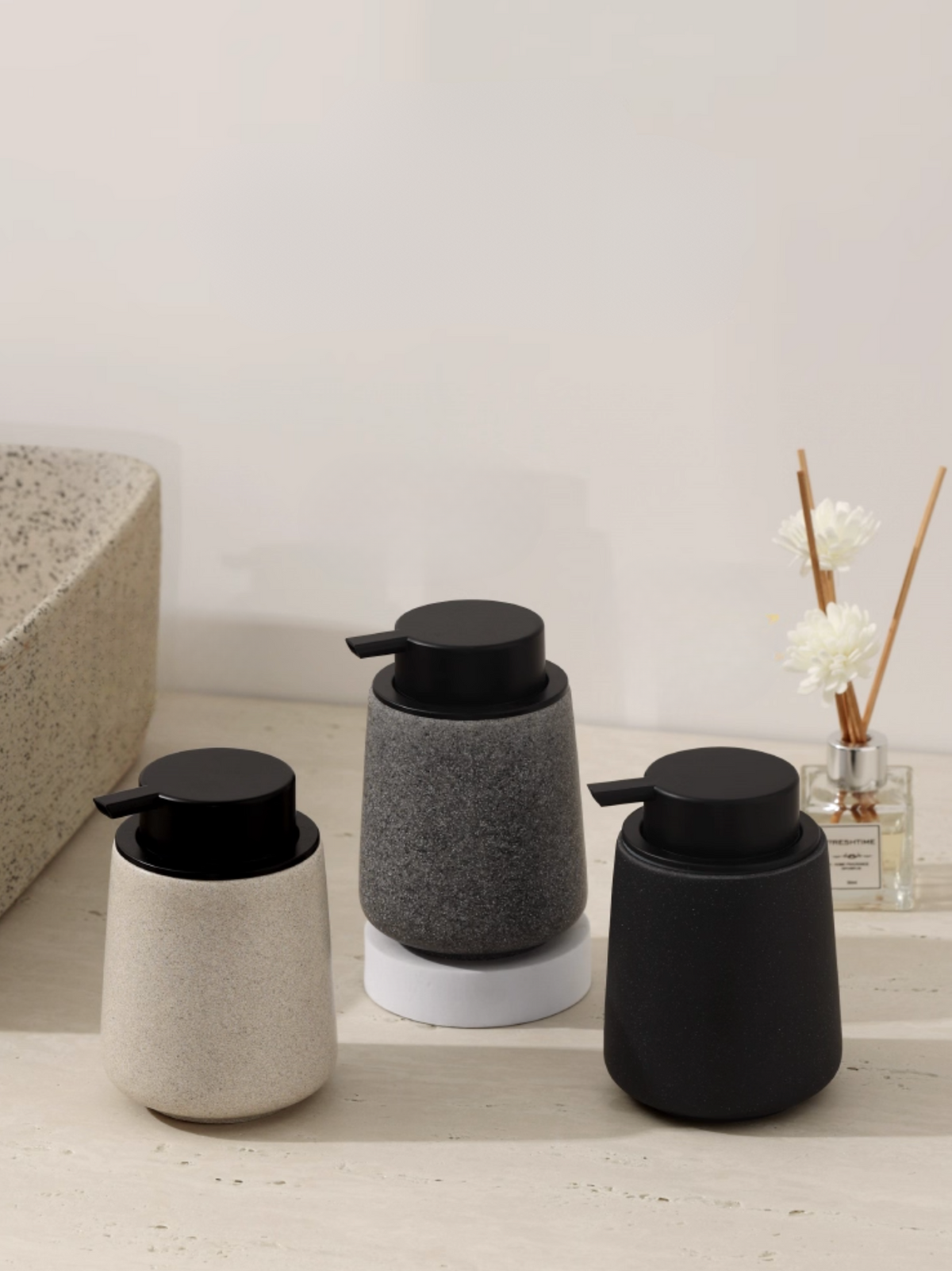 Three cylindrical soap dispensers in black, gray, and white on a bathroom counter.